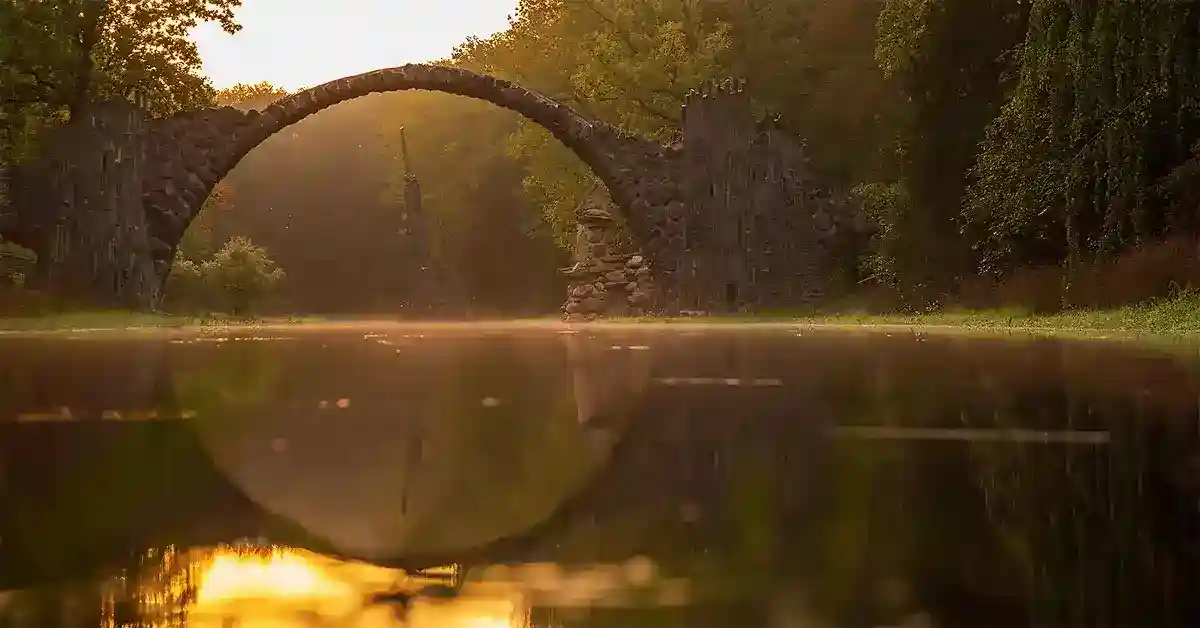 A stone arch bridge reflects on calm water during a foggy sunrise, surrounded by lush green trees.