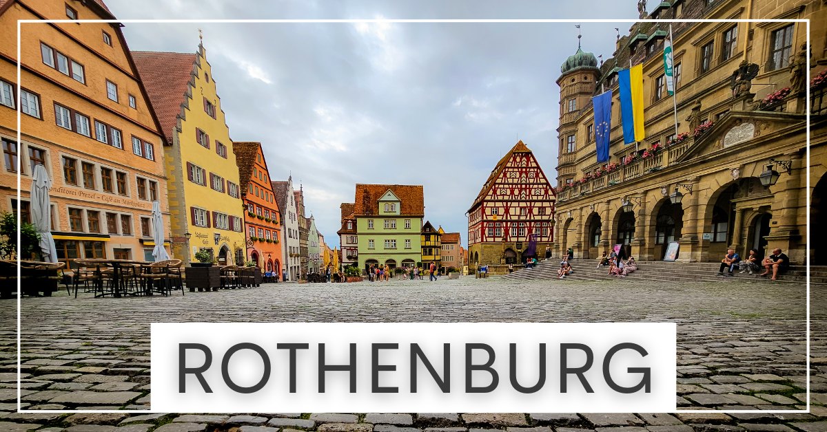 A cobblestone plaza in Rothenburg, Germany, surrounded by colorful historic buildings and a central, ornate building with a clock tower and flags.